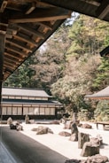 The image features a traditional Japanese rock garden surrounded by wooden buildings and lush greenery. The garden consists of carefully placed rocks on a bed of raked white gravel. The architecture includes wooden elements and shoji doors, with the surrounding trees providing a serene and natural backdrop.