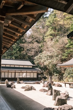 The image features a traditional Japanese rock garden surrounded by wooden buildings and lush greenery. The garden consists of carefully placed rocks on a bed of raked white gravel. The architecture includes wooden elements and shoji doors, with the surrounding trees providing a serene and natural backdrop.