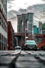 A striking view of the Brooklyn Bridge is captured from a low vantage point on a cobblestone street. The bridge's iconic arches and cables stand prominently against a cloudy sky. In the foreground, a black SUV and an ice cream truck are visible, adding a sense of urban life. On either side, buildings and bare trees frame the scene, while an American flag flutters in the middle distance.