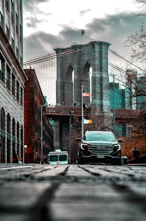 A striking view of the Brooklyn Bridge is captured from a low vantage point on a cobblestone street. The bridge's iconic arches and cables stand prominently against a cloudy sky. In the foreground, a black SUV and an ice cream truck are visible, adding a sense of urban life. On either side, buildings and bare trees frame the scene, while an American flag flutters in the middle distance.
