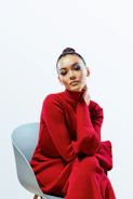 A person wearing a long red turtleneck dress sits on a minimalist chair with a light gray seat. The individual has a calm expression and their hand gently touches their neck. The background is a plain white, enhancing the vibrant red of the dress.