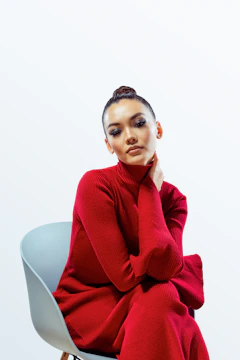 A person wearing a long red turtleneck dress sits on a minimalist chair with a light gray seat. The individual has a calm expression and their hand gently touches their neck. The background is a plain white, enhancing the vibrant red of the dress.