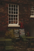 An outdoor scene with a brick wall featuring a vintage window showcasing a variety of violins. Below the window, there are several chalkboards displaying concert listings and events. A red cello leans against the wall, adding a splash of color. In the foreground, there are green metal chairs and tables, suggesting an outdoor seating area.