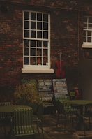 An outdoor scene with a brick wall featuring a vintage window showcasing a variety of violins. Below the window, there are several chalkboards displaying concert listings and events. A red cello leans against the wall, adding a splash of color. In the foreground, there are green metal chairs and tables, suggesting an outdoor seating area.