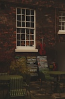 An outdoor scene with a brick wall featuring a vintage window showcasing a variety of violins. Below the window, there are several chalkboards displaying concert listings and events. A red cello leans against the wall, adding a splash of color. In the foreground, there are green metal chairs and tables, suggesting an outdoor seating area.