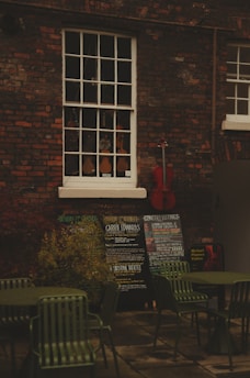 An outdoor scene with a brick wall featuring a vintage window showcasing a variety of violins. Below the window, there are several chalkboards displaying concert listings and events. A red cello leans against the wall, adding a splash of color. In the foreground, there are green metal chairs and tables, suggesting an outdoor seating area.