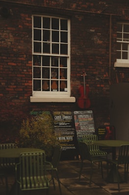 An outdoor scene with a brick wall featuring a vintage window showcasing a variety of violins. Below the window, there are several chalkboards displaying concert listings and events. A red cello leans against the wall, adding a splash of color. In the foreground, there are green metal chairs and tables, suggesting an outdoor seating area.