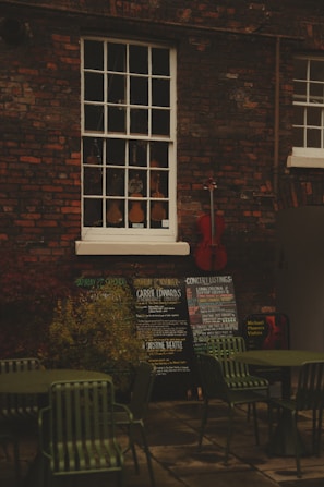 An outdoor scene with a brick wall featuring a vintage window showcasing a variety of violins. Below the window, there are several chalkboards displaying concert listings and events. A red cello leans against the wall, adding a splash of color. In the foreground, there are green metal chairs and tables, suggesting an outdoor seating area.
