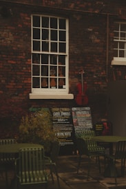 An outdoor scene with a brick wall featuring a vintage window showcasing a variety of violins. Below the window, there are several chalkboards displaying concert listings and events. A red cello leans against the wall, adding a splash of color. In the foreground, there are green metal chairs and tables, suggesting an outdoor seating area.