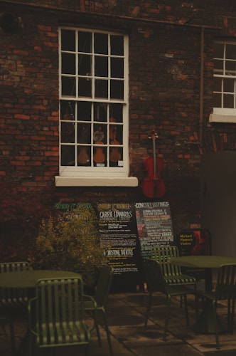 An outdoor scene with a brick wall featuring a vintage window showcasing a variety of violins. Below the window, there are several chalkboards displaying concert listings and events. A red cello leans against the wall, adding a splash of color. In the foreground, there are green metal chairs and tables, suggesting an outdoor seating area.