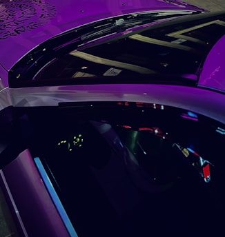A close-up view of a car with purple lighting reflecting on its exterior surfaces. The roof appears glossy with a decorative pattern, and there are subtle hints of an illuminated dashboard seen through the window. The lighting adds a futuristic and vibrant effect to the scene.