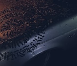 Calligraphic lettering covers the surface of a wet car hood, with droplets of water scattered across it. The lighting creates a contrast between the intricate black script and the metallic color of the car, reflecting a moody ambiance.