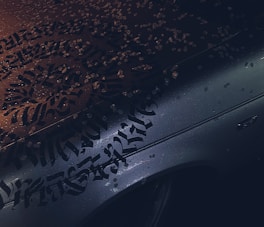 Calligraphic lettering covers the surface of a wet car hood, with droplets of water scattered across it. The lighting creates a contrast between the intricate black script and the metallic color of the car, reflecting a moody ambiance.