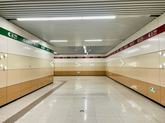 A clean, well-lit subway corridor with beige and white walls, featuring directional signage in green and red. The ceiling has a grid pattern with fluorescent lighting, and the floor is polished with marked guidance lines.