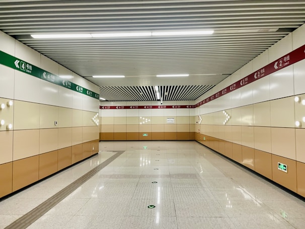 A clean, well-lit subway corridor with beige and white walls, featuring directional signage in green and red. The ceiling has a grid pattern with fluorescent lighting, and the floor is polished with marked guidance lines.