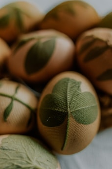 Eggs are decorated with green leaf patterns, likely created using a natural dyeing technique. The leaves appear to be pressed against the eggs, creating a detailed imprint of their veins and shapes. The colors are muted, with a soft focus that adds to an organic, rustic feel.