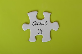 A white jigsaw puzzle piece with the words 'Contact Us' written on it is set against a bright lime green background.