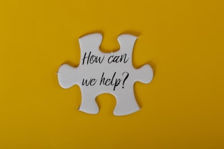A large white puzzle piece is centered against a bright yellow background. The text 'How can we help?' is written in black, elegant script on the puzzle piece.