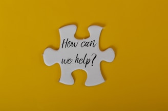 A large white puzzle piece is centered against a bright yellow background. The text 'How can we help?' is written in black, elegant script on the puzzle piece.