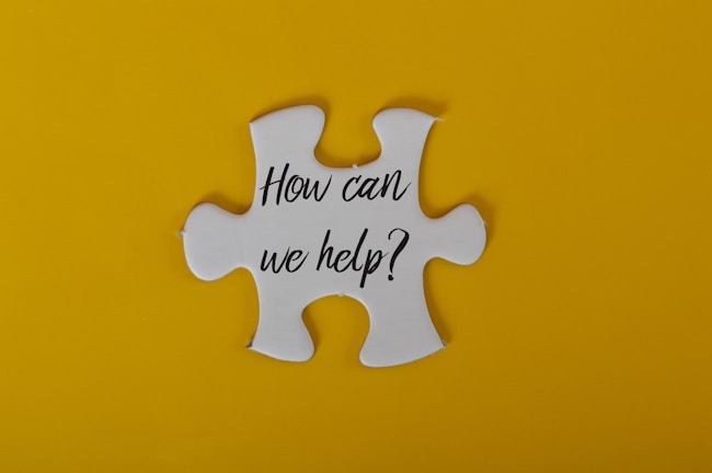A large white puzzle piece is centered against a bright yellow background. The text 'How can we help?' is written in black, elegant script on the puzzle piece.