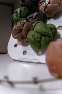 Several fabric bundles with decorative pins in green, brown, and dark tones are displayed on a white perforated stand. There is a pinkish-orange object in the foreground and blurred elements at the bottom.