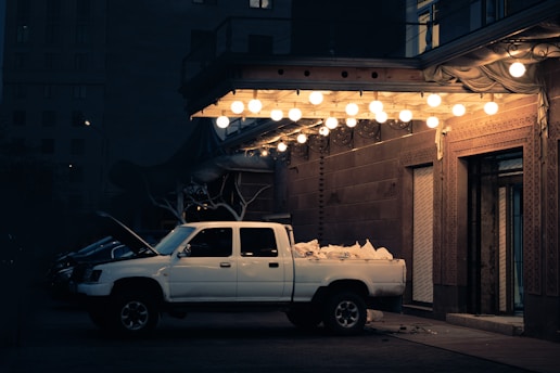 A white pickup truck is parked under a canopy lined with glowing bulbs. The truck's cargo bed is filled with bags. The hood is open, possibly indicating maintenance or a mechanical issue. The scene is set against a building with ornate details, and the lighting creates a warm contrast against the dark evening.