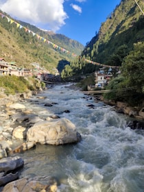 A flowing river with white water rapids runs through a picturesque valley surrounded by lush green hills. Colorful prayer flags are strung across the scene, and small structures line the riverbanks, showcasing a blend of natural beauty and human settlement. The sky is clear with scattered clouds, enhancing the serene yet dynamic atmosphere.