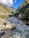 A flowing river with white water rapids runs through a picturesque valley surrounded by lush green hills. Colorful prayer flags are strung across the scene, and small structures line the riverbanks, showcasing a blend of natural beauty and human settlement. The sky is clear with scattered clouds, enhancing the serene yet dynamic atmosphere.