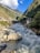 A flowing river with white water rapids runs through a picturesque valley surrounded by lush green hills. Colorful prayer flags are strung across the scene, and small structures line the riverbanks, showcasing a blend of natural beauty and human settlement. The sky is clear with scattered clouds, enhancing the serene yet dynamic atmosphere.