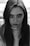 A black and white portrait of a young person with long hair, gazing directly at the viewer. The image focuses on the face and upper body, with a subtle expression. The lighting emphasizes facial features, shadows, and natural texture.