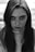 A black and white portrait of a young person with long hair, gazing directly at the viewer. The image focuses on the face and upper body, with a subtle expression. The lighting emphasizes facial features, shadows, and natural texture.