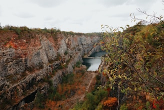 A scenic view of a deep quarry with steep rocky walls and a tranquil body of water at the bottom. Trees with autumn foliage frame the scene, adding a touch of greenery and reddish hues. A small bridge crosses over the water, connecting the two sides of the quarry.