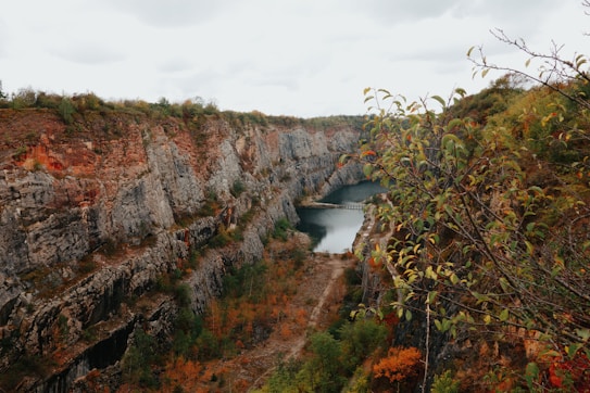 A scenic view of a deep quarry with steep rocky walls and a tranquil body of water at the bottom. Trees with autumn foliage frame the scene, adding a touch of greenery and reddish hues. A small bridge crosses over the water, connecting the two sides of the quarry.
