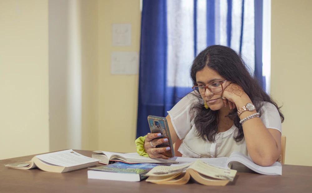 A student at a desk of open books, reading on her phone.