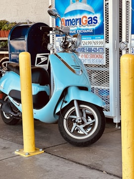 A light blue scooter is parked near a yellow bollard next to an AmeriGas propane exchange display. There is a blue mailbox and some potted plants in the background.