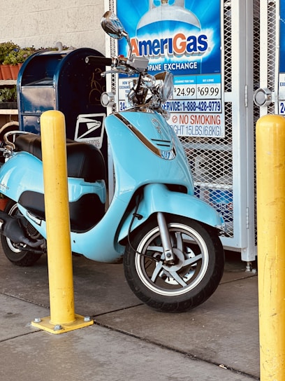 A light blue scooter is parked near a yellow bollard next to an AmeriGas propane exchange display. There is a blue mailbox and some potted plants in the background.