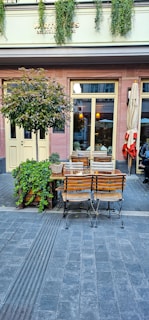A quaint outdoor café setting with wooden tables and chairs on a paved street. Lush green plants decorate the exterior, and vintage-style lanterns hang inside the building. The atmosphere suggests a cozy and inviting place to relax.