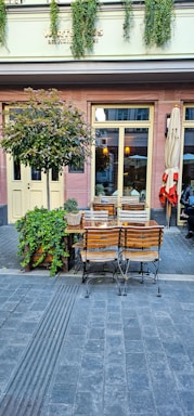 A quaint outdoor café setting with wooden tables and chairs on a paved street. Lush green plants decorate the exterior, and vintage-style lanterns hang inside the building. The atmosphere suggests a cozy and inviting place to relax.
