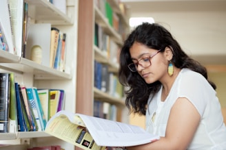 A person with glasses and colorful earrings is reading a book in a library. Shelves filled with various books are visible in the background, creating a scholarly setting.
