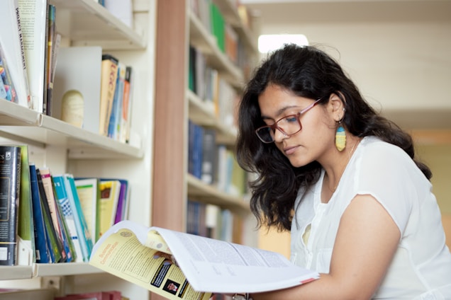 A person with glasses and colorful earrings is reading a book in a library. Shelves filled with various books are visible in the background, creating a scholarly setting.