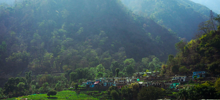 A small village is nestled among lush green terraces on the hillsides. The area is surrounded by dense forests and towering mountains, creating a tranquil and scenic landscape. The buildings in the village are of varying colors, adding vibrancy to the setting.