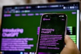 A hand holding a smartphone displaying an introduction screen for ChatGPT with the text visible. The background features a blurred computer screen showing similar content, creating a mirror effect. Both screens have a green and purple color scheme.