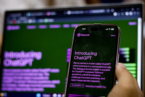 A hand holding a smartphone displaying an introduction screen for ChatGPT with the text visible. The background features a blurred computer screen showing similar content, creating a mirror effect. Both screens have a green and purple color scheme.