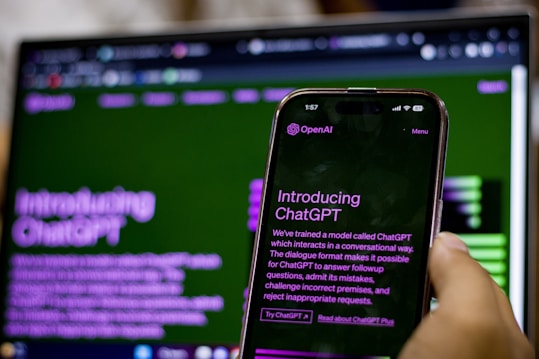 A hand holding a smartphone displaying an introduction screen for ChatGPT with the text visible. The background features a blurred computer screen showing similar content, creating a mirror effect. Both screens have a green and purple color scheme.
