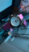 A wristwatch with a round face and pink metallic strap is positioned on a reflective surface. The watch has a white dial with elegant text and a sleek design that gives a luxurious appearance. Lighting and shadows create an artistic and high-contrast effect, highlighting the watch.