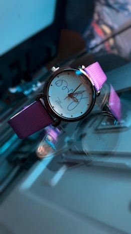 A wristwatch with a round face and pink metallic strap is positioned on a reflective surface. The watch has a white dial with elegant text and a sleek design that gives a luxurious appearance. Lighting and shadows create an artistic and high-contrast effect, highlighting the watch.