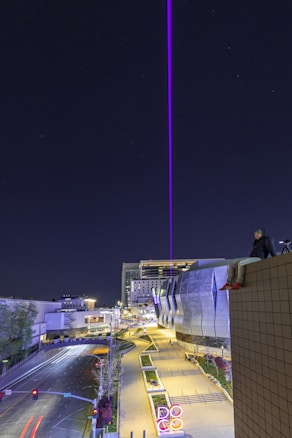 A cityscape at night featuring a bright purple laser beam extending vertically into the sky. The scene includes modern architecture with a large, metallic-sided building and an illuminated walkway. A person is sitting on the edge of a high structure, possibly taking photographs, with a tripod next to them. The roads below are empty, and artificial lights highlight various structures.
