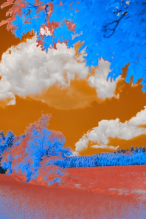 The image features a surreal landscape with digitally altered colors. Trees and foliage are depicted in vibrant blue and orange hues. The sky is a rich orange, contrasted by white clouds. The ground appears in a combination of blue and orange tones, adding to the dreamlike quality of the scene.