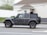 A grey Jeep Rubicon is driving on a suburban street, with a blurred background indicating motion. The vehicle has large tires and a spare tire mounted on the back. A person is visible through the window.