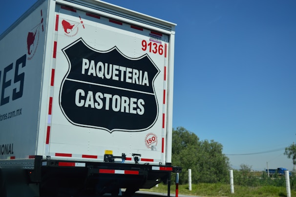 The rear view of a delivery truck with the label 'PAQUETERIA CASTORES' displayed prominently on a black emblem on the back door. The truck has the number '9136' and an 'ISO' certification badge. The background shows a clear blue sky and some greenery.